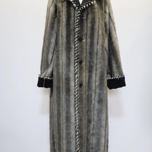 May include: A long, gray and black faux fur coat with vertical stripes. The coat features a black collar and cuffs, and a decorative stitched trim down the front and around the collar. The coat is buttoned up and displayed on a mannequin.