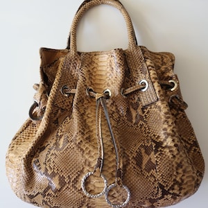 May include: A brown and tan snakeskin handbag with a drawstring closure and a top handle. The bag features silver-toned metal hardware, including rings and grommets. The snakeskin pattern is visible across the entire bag.