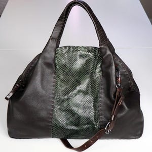 Pode incluir: Uma mala de mão de couro castanho escuro com um painel de pele de cobra. A mala tem uma pega superior e uma alça de ombro removível. O painel de pele de cobra é verde e cinzento. A mala tem um fecho de correr e um fecho de metal.
