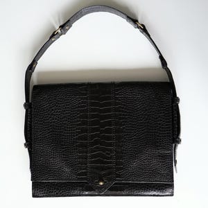 Puede incluir: Bolso de mano de cuero negro con un patrón texturizado similar al de un cocodrilo. El bolso tiene una correa larga y ajustable y un cierre de solapa con un broche de metal. El diseño del bolso es rectangular con una costura vertical en el centro.