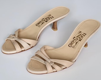 May include: A pair of beige leather Salvatore Ferragamo heeled sandals. The open-toe shoes feature a knotted strap design and a small wooden heel. The brand name is printed on the insole.