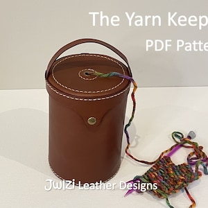 May include: A brown leather yarn keeper with a handle and a white stitch detail. The yarn keeper is filled with colorful yarn and is sitting on a white surface. The text "The Yarn Keeper PDF Pattern" is above the yarn keeper. The text "Julzi Leather Designs" is below the yarn keeper.