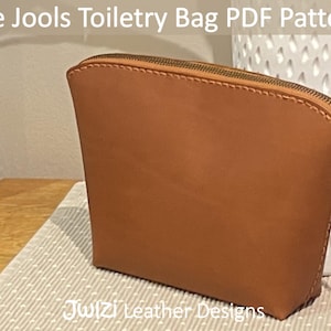 Peut inclure: Une trousse de toilette en cuir marron avec une fermeture éclair. La trousse est posée sur une surface blanche et marron. Le texte "The Jools Toiletry Bag PDF Pattern" est en haut de l'image. Le texte "4 sizes!" est à gauche de l'image. Le texte "Julzi Leather Designs" est en bas de l'image.