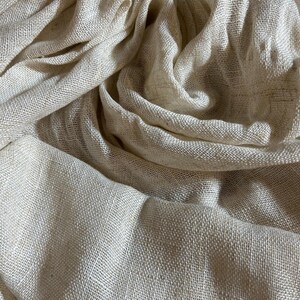 May include: Close-up of a beige linen fabric with a textured weave. The fabric is draped and folded, creating shadows and highlights. The material appears soft and lightweight, suitable for clothing or home decor.