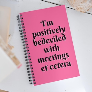 Puede incluir: Un cuaderno rosa con espiral con el texto "I'm positively bedeviled with meetings et cetera" en negro. El cuaderno está abierto, mostrando la portada. El fondo es una superficie blanca.