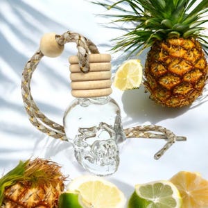 Puede incluir: Un ambientador para coche con forma de calavera de cristal transparente con tapón de madera y cuerda trenzada. El ambientador está colgado, con una piña, limón y lima de fondo. El ambientador es un artículo decorativo.