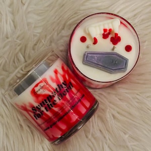 May include: Two glass candles with white wax and red splatter designs. One candle has the text "Sympathy for the Devil". The other candle has a coffin and vampire teeth decorations.