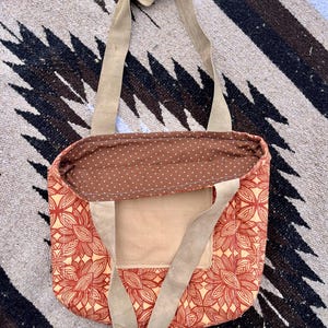 May include: A large, red and cream tote bag with a floral pattern. The bag has two long, brown straps and a small, brown pocket on the front.
