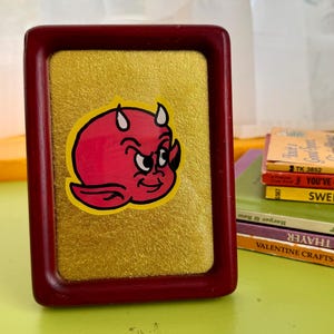 May include: A framed picture featuring a red devil cartoon character with horns and a mischievous expression. The frame is a dark red color, and the background is gold. The picture is on a green surface.