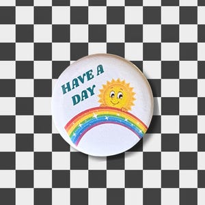 May include: A white button with the text "HAVE A DAY" in teal. The button features a smiling yellow sun over a rainbow. The background is a black and white checkered pattern.
