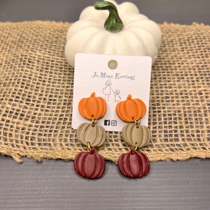 Puede incluir: Un par de pendientes colgantes con tres abalorios de calabaza apilados. La calabaza superior es naranja, la calabaza del medio es verde apagado y la calabaza inferior es burdeos. Los pendientes están en una tarjeta blanca con el texto "Jo Mama Earrings" y los iconos de las redes sociales.