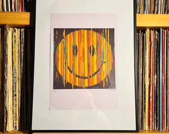 Puede incluir: Una impresión enmarcada que presenta una cara sonriente amarilla compuesta por rayas verticales multicolores en tonos naranja, rojo y verde sobre un fondo oscuro. La impresión se muestra sobre un letrero de madera con el texto "NOW PLAYING".