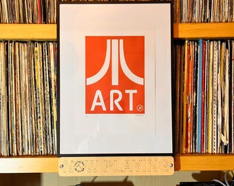 Puede incluir: Una impresión enmarcada que presenta un logotipo de Atari blanco y la palabra ART en blanco sobre un fondo rectangular naranja brillante. Debajo del marco, hay un letrero de madera clara con el texto NOW PLAYING, que incluye un pequeño logotipo circular.
