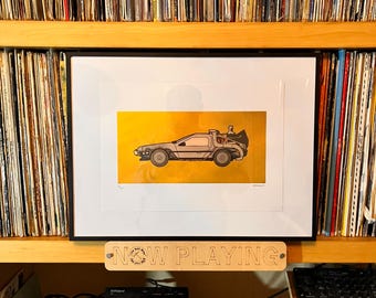 Puede incluir: Una lámina artística enmarcada que muestra una ilustración de perfil de un coche deportivo plateado sobre un fondo amarillo sólido. La lámina está en un marco negro y se sitúa sobre un letrero de madera con el texto NOW PLAYING grabado.