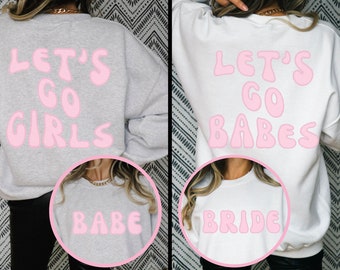 May include: Two gray and white sweatshirts with pink text. The first sweatshirt says "Let's Go Girls" and the second sweatshirt says "Let's Go Babes". A smaller gray sweatshirt with pink text that says "Babe" is in front of the first sweatshirt. A smaller white sweatshirt with pink text that says "Bride" is in front of the second sweatshirt.