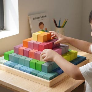 Puede incluir: Un juego de bloques de madera de colores del arcoíris dispuestos en forma de pirámide sobre una bandeja de madera. Los bloques son de varios tonos de azul, verde, amarillo, naranja y rojo. El juego está sobre una mesa de madera clara.