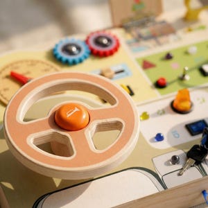 May include: Wooden toy car dashboard with a steering wheel, horn button, and various interactive elements. The steering wheel is light brown with an orange center. Other features include colorful dials, switches, and a key. The toy is designed for children.