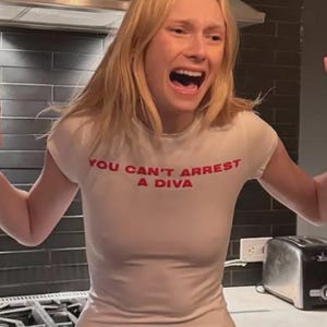 May include: A short-sleeved, cream-colored fitted t-shirt featuring the text "YOU CAN'T ARREST A DIVA" printed in bold red capital letters across the chest. The garment has a simple crew neckline and a slim, form-fitting silhouette.