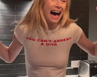 Op de afbeelding: Een crèmekleurig, nauwsluitend T-shirt met korte mouwen en de rode tekst "YOU CAN'T ARREST A DIVA" op de borst gedrukt. Het kledingstuk heeft een eenvoudige ronde hals en een slank, nauwsluitend silhouet.