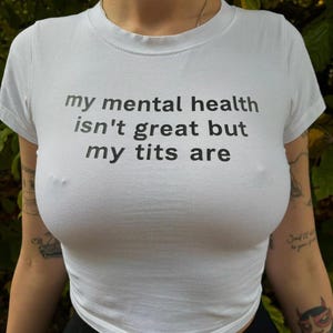 May include: White, short-sleeved t-shirt with the text "my mental health isn't great but my tits are" printed in black. The shirt is a fitted style.