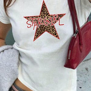 May include: White t-shirt featuring a red-outlined star with a leopard print design. The word "STARGIRL" is printed in red across the star. A red purse hangs on the right side.