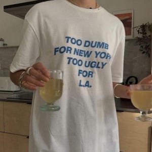Może przedstawiać: Biały t-shirt z krótkim rękawem z niebieskim napisem "TOO DUMB FOR NEW YORK TOO UGLY FOR L.A." w połączeniu z niebieskimi szortami sportowymi z białym logo na lewej nogawce. Koszulka ma okrągły dekolt i luźny, swobodny krój.