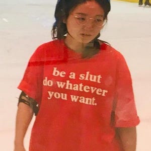 Puede incluir: Una camiseta roja de manga corta con el texto impreso en blanco 'be a slut do whatever you want.' La prenda se lleva con un casco protector azul brillante y gafas de montura fina en una pista de patinaje sobre hielo cubierta y luminosa.