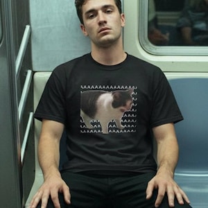 Puede incluir: Una camiseta negra de manga corta con un gráfico de un gato blanco y negro en el centro. El diseño incluye la letra A repetida en un patrón triangular por encima y por debajo del gato, creando un elemento visual muy distintivo.