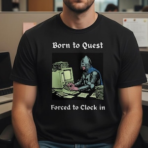 May include: Black t-shirt with the text "Born to Quest, Forced to Clock in." The shirt features a graphic of a knight at a computer. The person in the image is wearing the shirt.