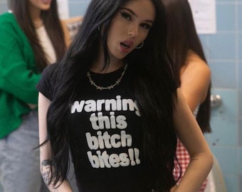 Op de afbeelding: Een zwart kortemouwen crop top met de witte tekst "warning this bitch bites!!" op de voorkant gedrukt. Het kledingstuk heeft een ronde hals en wordt gecombineerd met een zilverkleurige ketting met kleine stekels.