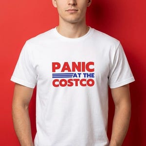 Könnte beinhalten: Weißes T-Shirt mit dem Text "PANIC AT THE COSTCO" in Rot und Blau. Das Shirt hat einen Rundhalsausschnitt und kurze Ärmel. Der Text ist mittig auf der Vorderseite des Shirts platziert.