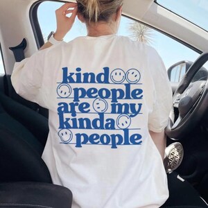 Pode incluir: Uma t-shirt branca com texto azul nas costas que diz "kind people are my kinda people" disposto em cinco linhas horizontais. O design inclui pequenos ícones de rostos sorridentes azuis intercalados entre as palavras na t-shirt.