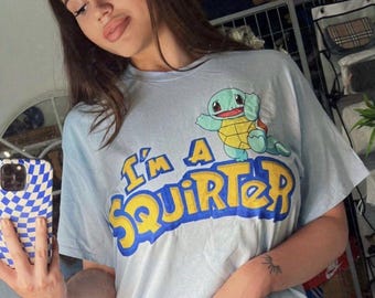 May include: A light blue short-sleeved t-shirt featuring a graphic of the blue and yellow turtle character Squirtle positioned above the yellow and blue text that reads I'm A SQUIRTER in a bold, rounded, cartoon-style font.