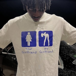 以下が含まれることがあります： 青いグラフィックと「Your Girlfriend」と「My Girlfriend」の文字が入った白いTシャツ。Tシャツはクルーネックで半袖です。