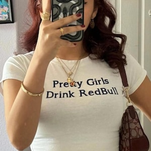 May include: A white short-sleeved t-shirt featuring the black text 'Pretty Girls Drink RedBull' printed on the chest. The shirt is worn with a brown patterned shoulder bag, a gold bangle, and layered gold necklaces.