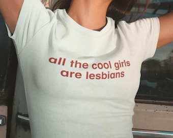 May include: A white short-sleeved t-shirt featuring the text "all the cool girls are lesbians" printed in a dark red, sans-serif font across the chest. The garment has a crew neckline and a fitted silhouette.