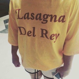 Może przedstawiać: Jaskrawożółty t-shirt z krótkim rękawem z ciemnobrązowym napisem Lasagna Del Rey nadrukowanym na piersi kursywą. Odzież ma luźny krój z szerokimi rękawami i jest zestawiona z białymi szortami z czarnymi wykończeniami.
