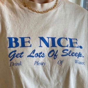 Peut inclure: T-shirt crème avec le texte bleu "BE NICE. Get Lots Of Sleep. Drink Plenty Of Water." Un collier chaîne doré est porté autour du cou. Le t-shirt a un col rond et des manches courtes.