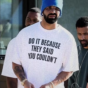 May include: A white short-sleeved t-shirt with a speckled texture and black text that reads DO IT BECAUSE THEY SAID YOU COULDN'T. The shirt is worn with a bright blue beanie cap, a gold wristwatch, and a gold bracelet on the wrist.