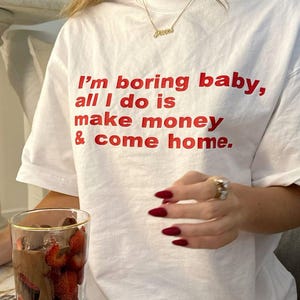 Może przedstawiać: Biały t-shirt z krótkim rękawem z czerwonym napisem "I'm boring baby, all I do is make money & come home." Koszulka ma okrągły dekolt i luźny krój, z wyraźnym nadrukiem graficznym umieszczonym na wysokości klatki piersiowej.