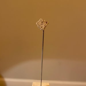 May include: A small, irregularly shaped, light brown rock is mounted on a thin metal rod. The rod is attached to a small, square, wooden base.