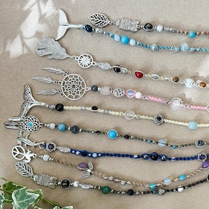 Op de afbeelding: Een verzameling kleurrijke kralenkettingen met verschillende bedels, waaronder een walvisstaart, een droomvanger en een vredesteken. De kettingen zijn in een regenboogpatroon op een bruin oppervlak gerangschikt.