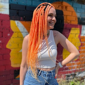Pode incluir: Uma mulher com tran&ccedil;as longas, de cor laranja e amarela brilhante, em frente a uma parede de tijolos colorida. Ela est&aacute; vestindo um top branco sem mangas e uma saia jeans.