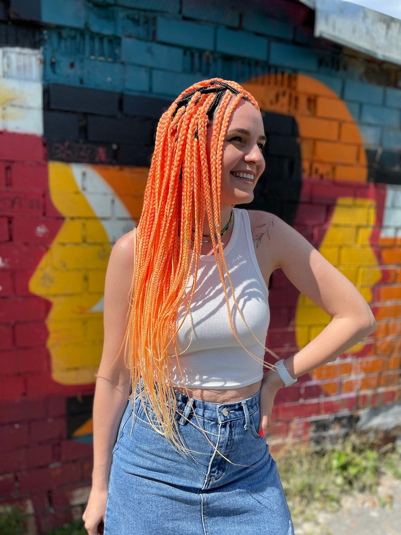 Pode incluir: Uma mulher com tran&ccedil;as longas, de cor laranja e amarela brilhante, em frente a uma parede de tijolos colorida. Ela est&aacute; vestindo um top branco sem mangas e uma saia jeans.