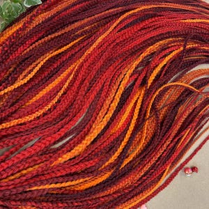 May include: A close-up of a bundle of red, orange, and brown braided yarn. The yarn is arranged in a wavy pattern, creating a textured and colorful display.