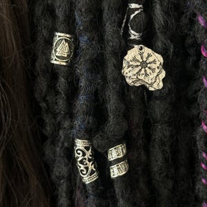 May include: Silver dreadlock beads with various designs, including a snowflake, a triangle, and a swirl pattern. The beads are attached to black dreadlocks.