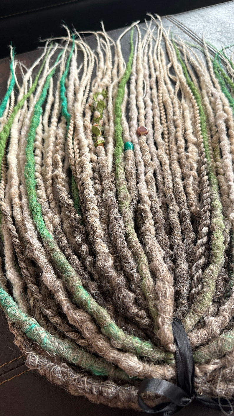 May include: A bundle of synthetic dreadlocks in shades of gray, white, and green. The dreadlocks are tied together with a black ribbon.