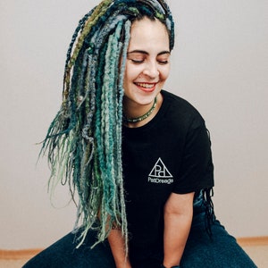 Pode incluir: Uma pessoa com dreadlocks longos, azuis e verdes est&aacute; vestindo uma camiseta preta com um logotipo branco em forma de tri&acirc;ngulo que diz "PattiDreadz". Ela est&aacute; vestindo jeans azuis e um rel&oacute;gio preto.