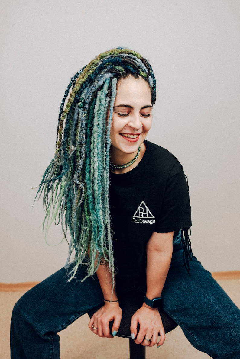 Pode incluir: Uma pessoa com dreadlocks longos, azuis e verdes est&aacute; vestindo uma camiseta preta com um logotipo branco em forma de tri&acirc;ngulo que diz "PattiDreadz". Ela est&aacute; vestindo jeans azuis e um rel&oacute;gio preto.
