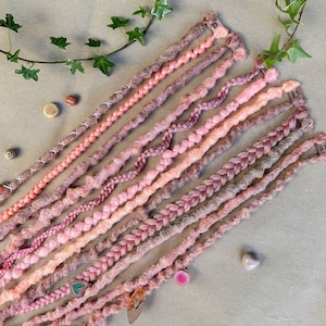 Peut inclure: Une collection de dreadlocks synthétiques de couleur rose et pêche avec différentes textures, notamment lisses, ondulées et tressées. Les dreadlocks sont disposées en éventail sur une surface brune.